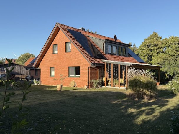 Von der Sonne erstrahltes EFH mit neuer PV-Anlage Rotes Haus mit Satteldach, Garten und Solarplatten, umgeben von Bäumen.