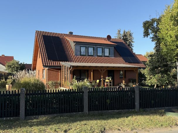 Südseite mit neuer PV-Anlage Holzhaus mit rotem Dach, Solarpanelen, Garten und Zaun. Klarer Himmel im Hintergrund.